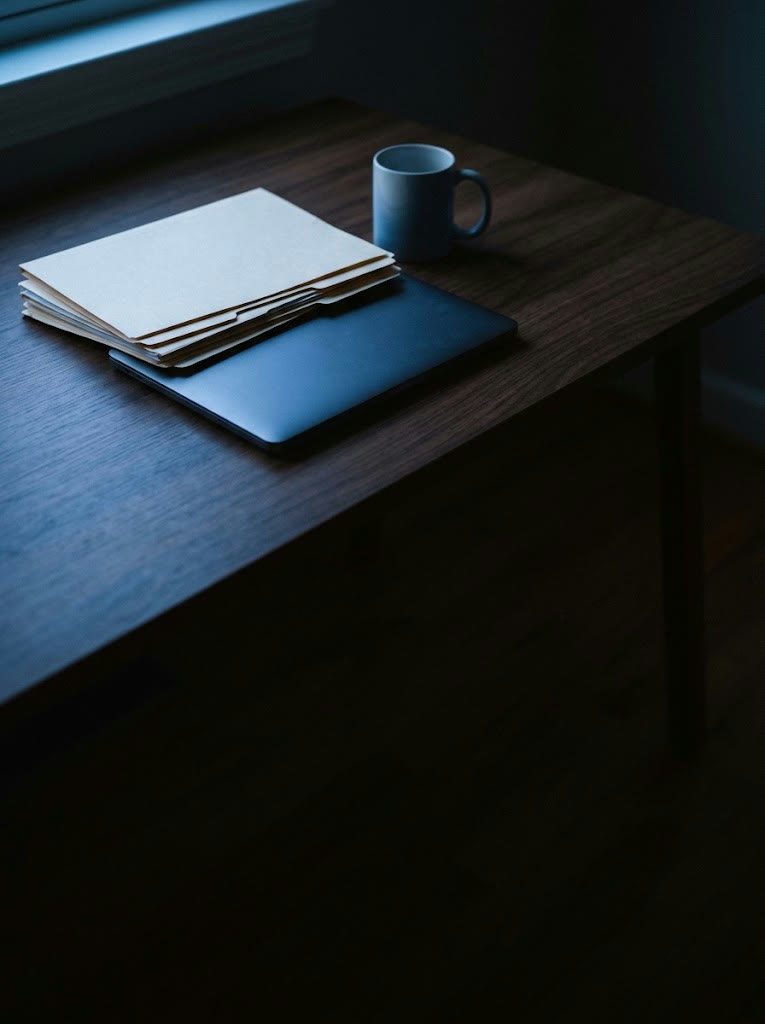 A stack of manila folders on a closed laptop beside a mug, on a desk in blue window light
